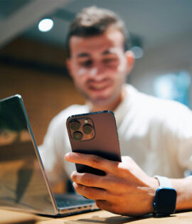 A person sits at a table, holding a smartphone in one hand and working on a laptop.