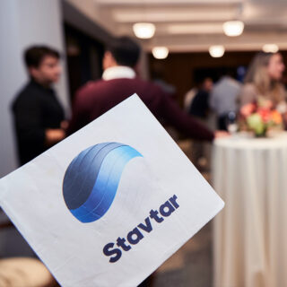 A napkin with the Stavtar logo in the foreground. People socialize at an indoor event space in the background.