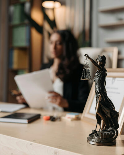 Bronze Lady Justice statue on desk with woman in background reviewing documents.