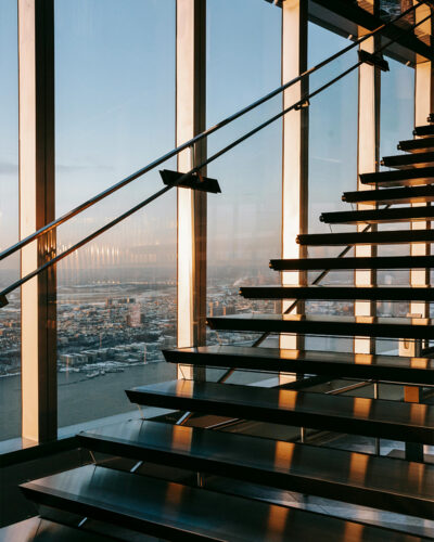 Modern glass staircase inside a skyscraper, with warm sunlight streaming through large windows, revealing a panoramic cityscape in the distance.