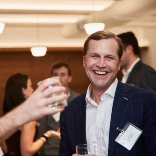 A man in a suit laughs while holding a drink at a social gathering.