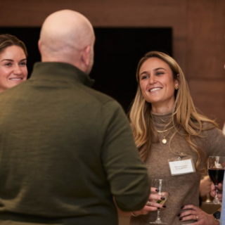 Woman, wearing a tan turtleneck and a name tag, holds a champagne flute and smiles at a man in a blue striped shirt holding a glass of red wine.